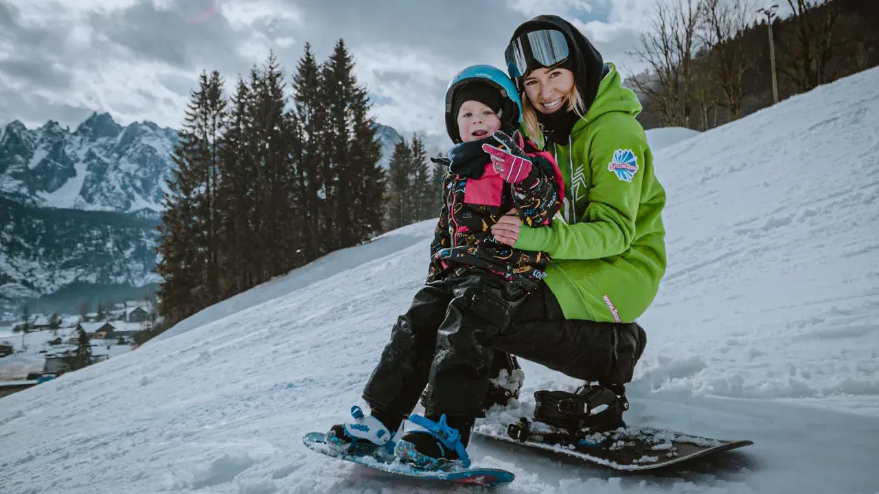 Uśmiechnięta instruktorka na stoku narciarskim dla dzieci trzyma małego snowboardzistę.