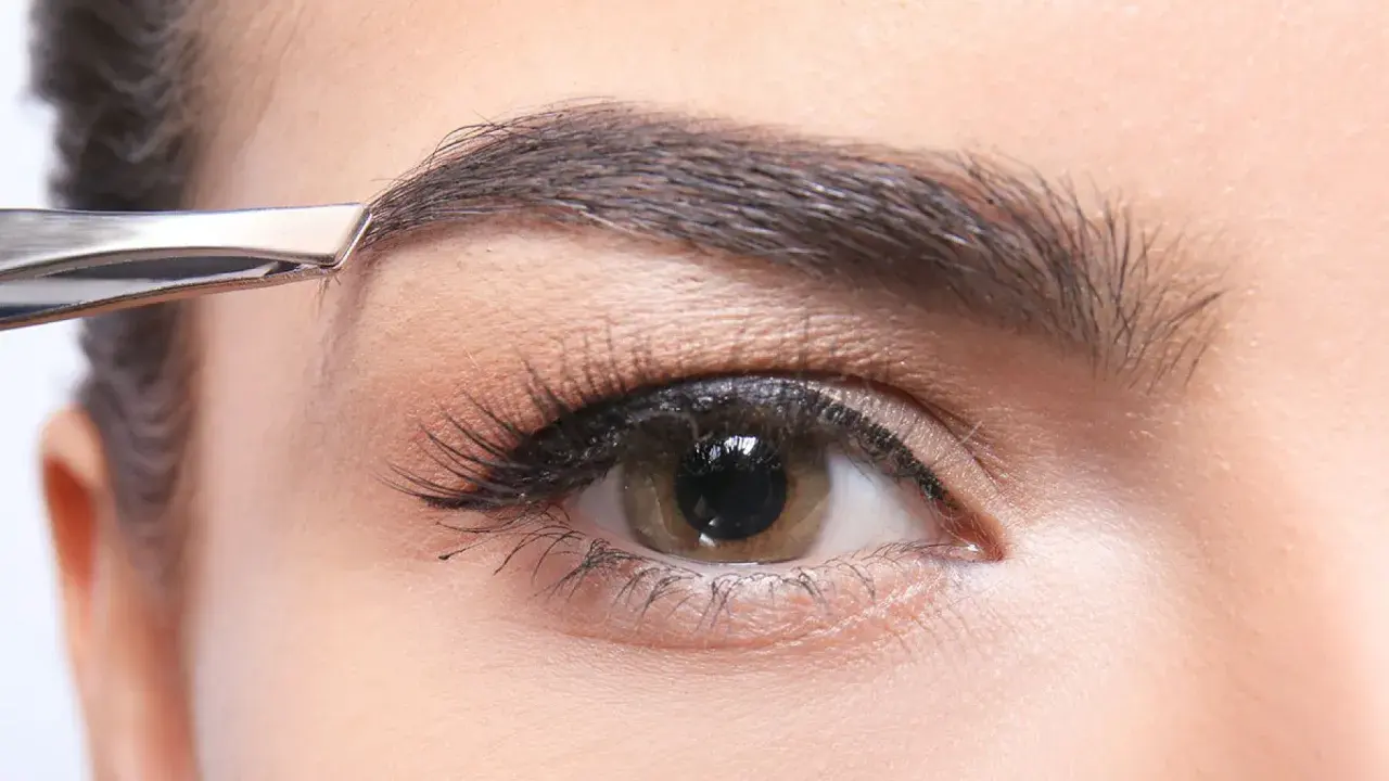 Une pince à épiler retire délicatement un poil du sourcil, affinant la ligne pour un regard parfait.