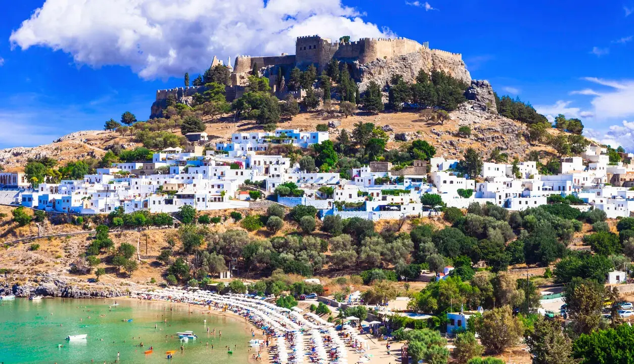 En vitkalkad by klättrar uppför en kulle mot en borgruin. Nedanför breder en strand ut sig med solstolar och turkost vatten. En vy över Rhodos karta Europa.
