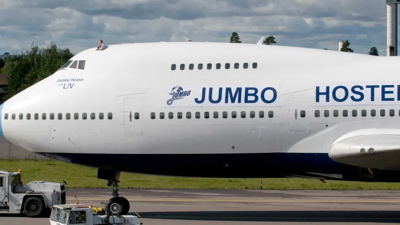 En person sitter på taket av ett jumbo stay. Flygplanet är vitt med blå detaljer och texten "JUMBO HOSTEL" syns tydligt.