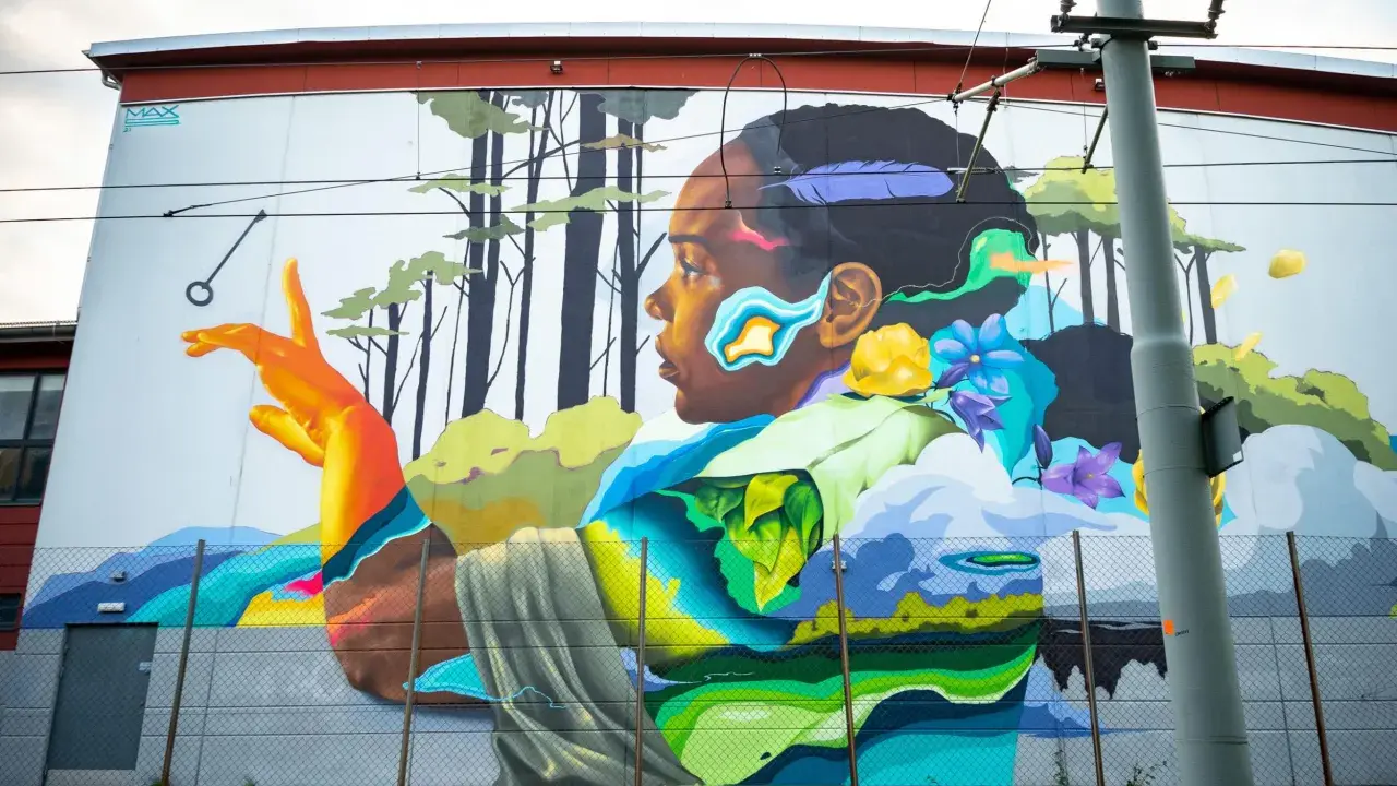 Färgstark street art på en vägg visar en person i profil med en fjäder i håret, omgiven av träd och blommor.