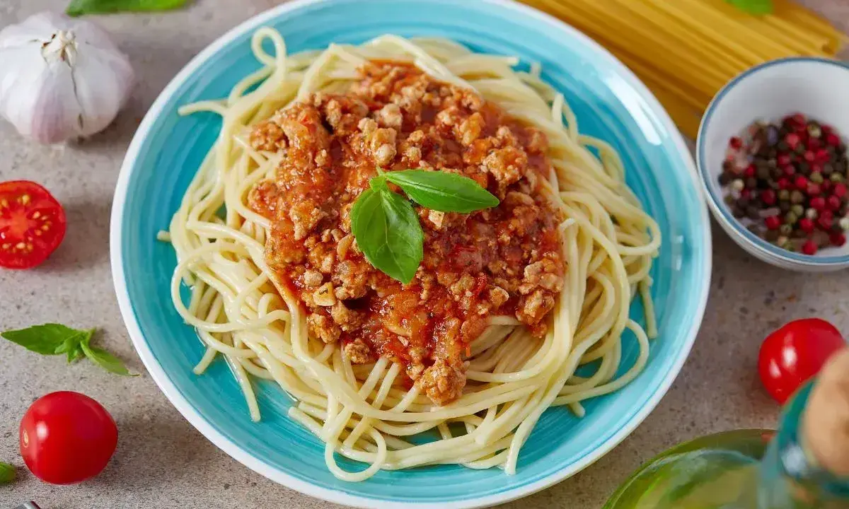 Pyszne spaghetti z bogatym sosem bolognese, udekorowane listkiem bazylii. Obok czosnek, pomidorki i przyprawy.