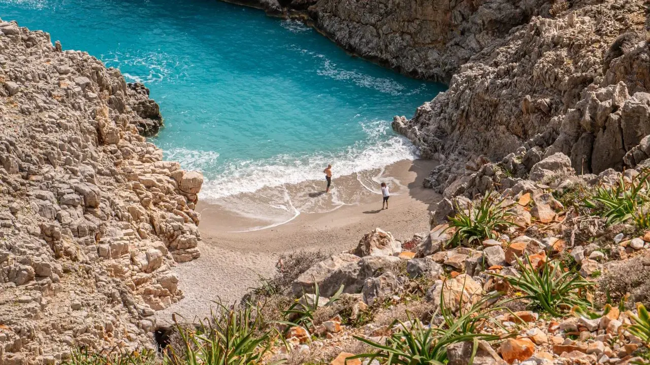 Malownicza zatoka z turkusową wodą i piaszczystą plażą, otoczona skalistymi klifami. Dwie osoby spacerują po brzegu. Chania plaże zachwycają.