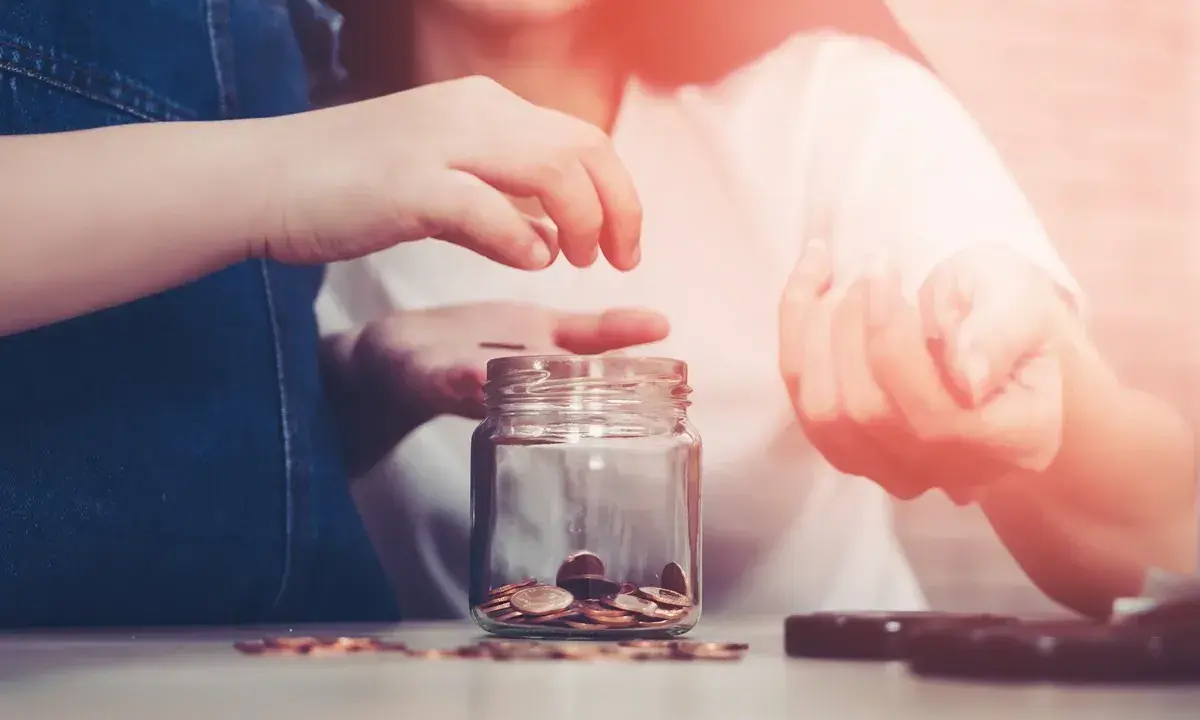Dwie osoby wrzucają monety do słoika, oszczędzając na przyszłość. Zastanawiasz się, ile wynoszą alimenty na studenta?