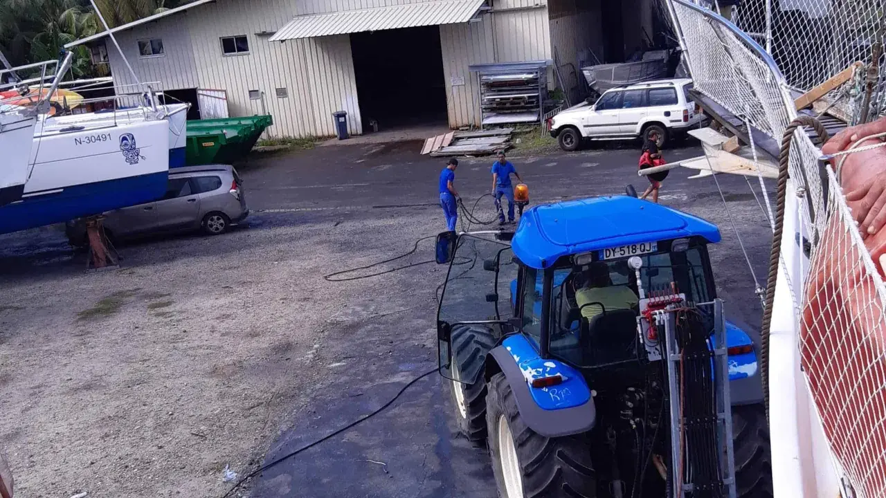 Tracteur bleu près d'un bateau, peut-être pour le prix d'entretien d'un bateau de 7m. Des ouvriers travaillent.