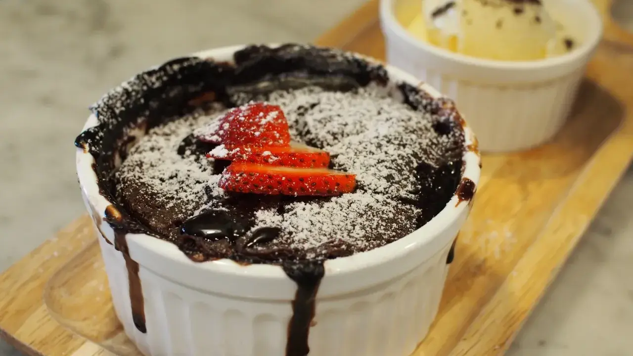 Pyszny, gorący lava cake z mikrofali, posypany cukrem pudrem i udekorowany truskawkami. Obok porcja lodów waniliowych.