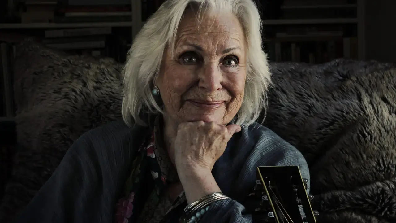 Äldre kvinna med grått hår, gitarr och en fluffig pläd. Trots att Marie Bergman är sjuk, lyser hennes ögon av liv.