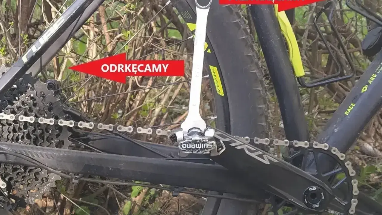 Klucz do pedałów rowerowych przy korbie, z czerwonymi strzałkami wskazującymi "Odkręcamy" i "Przykręcamy".