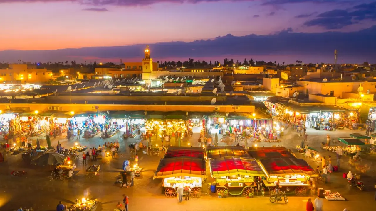 Der belebte marrakesch marktplatz erwacht bei Dämmerung zum Leben. Stände mit bunten Waren, Essensverkäufer und Menschenmassen beleben die Szene unter dem lila Himmel.