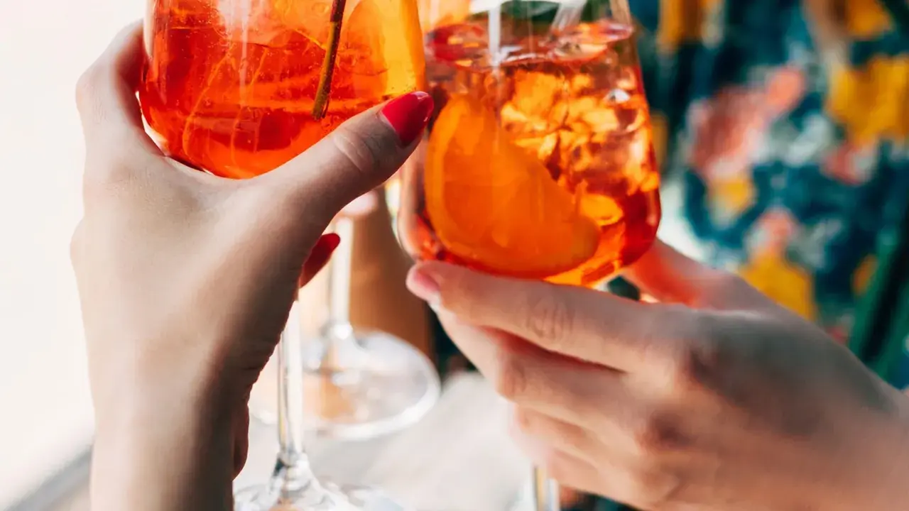 Zwei Hände halten Aperol Spritz Gläser mit Eis und Orangenscheiben. Ein perfektes Aperol Rezept für den Sommer.