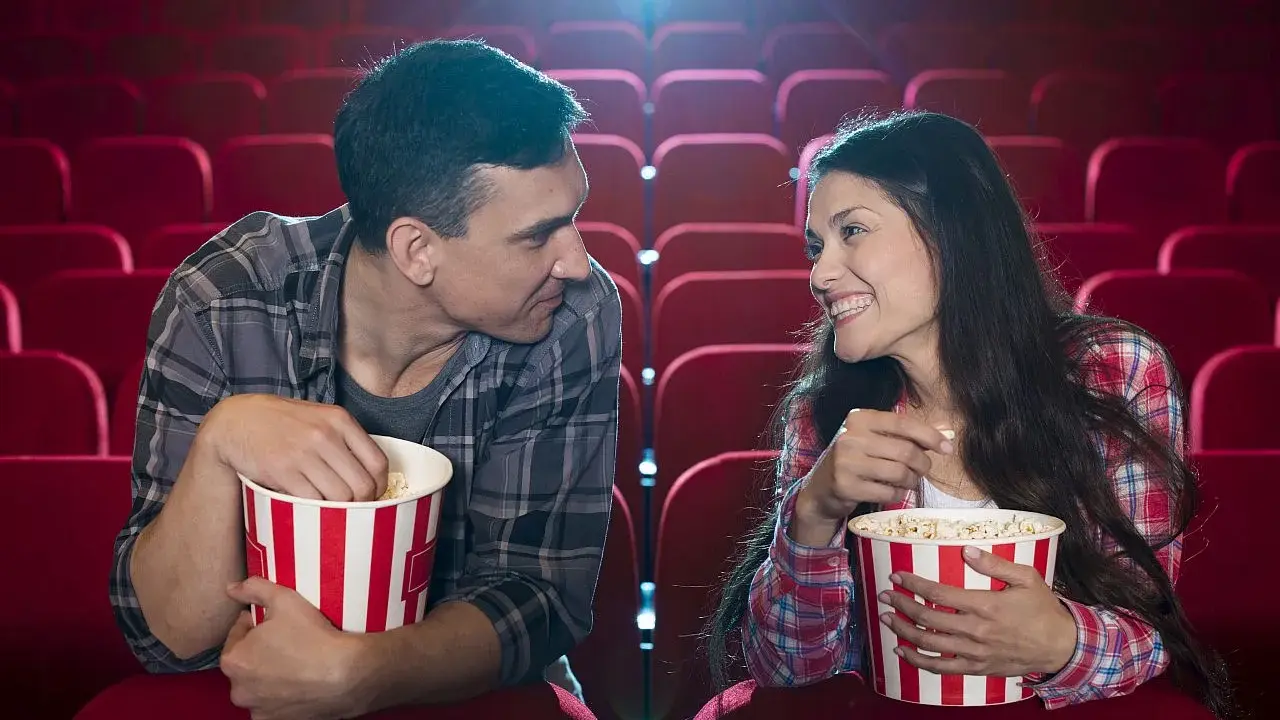 Para w kinie, zajadając popcorn, patrzy na siebie z uśmiechem. Ich spojrzenia sugerują, że to nie tylko film ich interesuje, ale też wzajemna bliskość.
