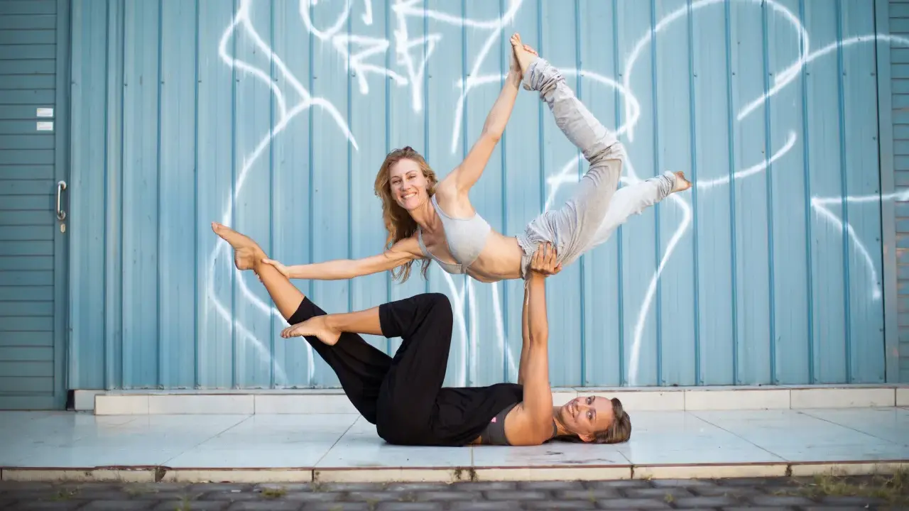 Två kvinnor utför acro yoga framför en blå vägg med graffiti. Den ena ligger på rygg och lyfter den andra som är i en böjd position.