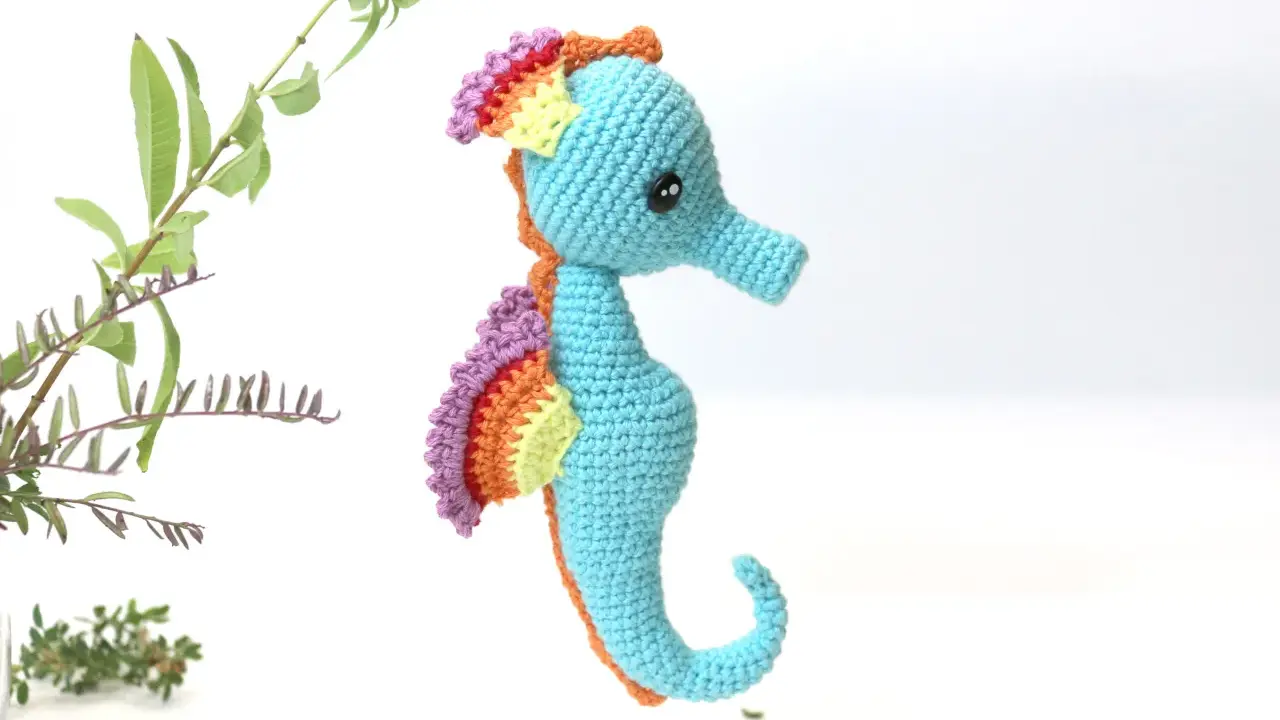 Caballito de mar amigurumi patron gratis. Un adorable caballito de mar tejido a crochet, con aletas arcoíris y cuerpo azul, listo para navegar en tu imaginación.
