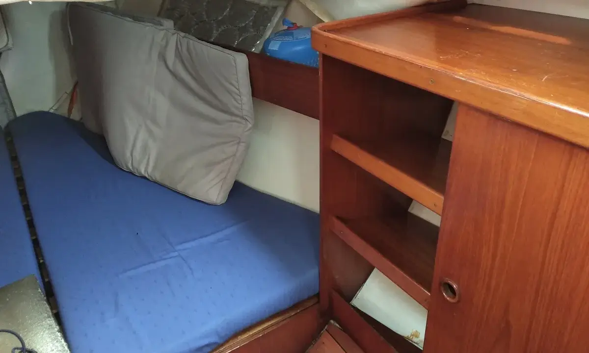 L'intérieur d'un voilier avec une couchette bleue, un coussin gris et des étagères en bois.