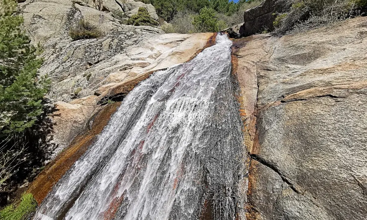 El agua desciende por la roca en la cascada del Hornillo, rodeada de vegetación y rocas graníticas bajo un cielo azul.