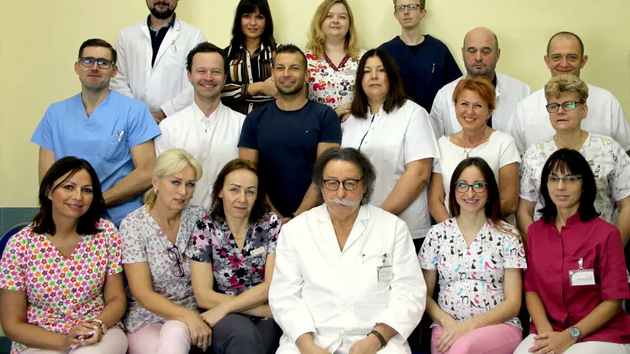 Grupa lekarzy i pielęgniarek w białych fartuchach i kolorowych strojach medycznych, pozująca do zdjęcia.