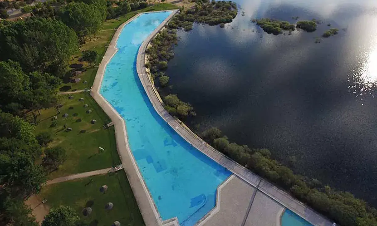 Vista aérea de la piscina natural de Riosequillo, con su agua azul brillante bordeada por césped verde y un lago oscuro.