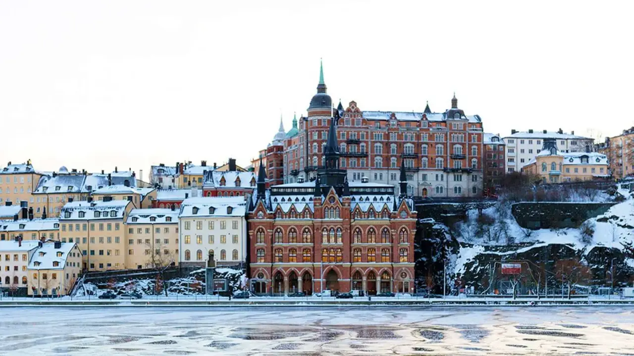 Vinter i Stockholm med snöklädda tak och isig vattenyta. Vackra byggnader reser sig mot en klar himmel.