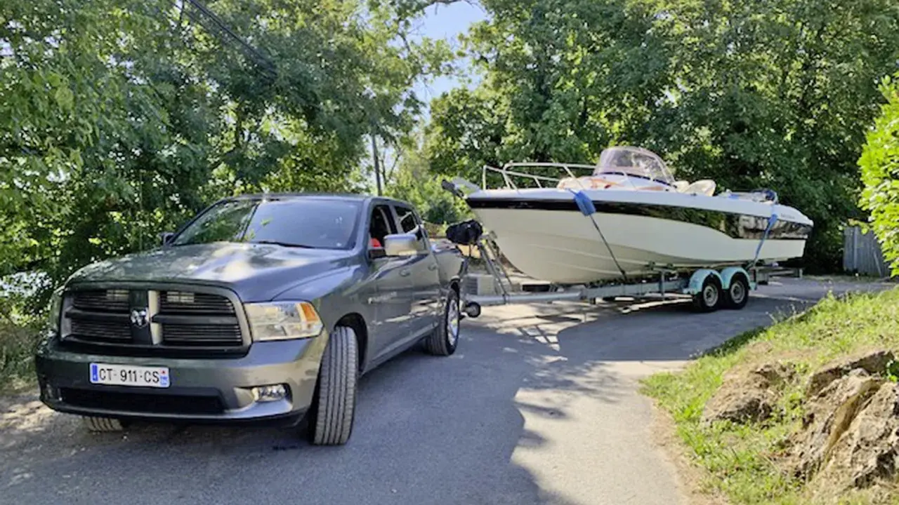 Un pick-up gris est prêt à tracter un bateau de 7m sur sa remorque, sous un ciel bleu et entouré de verdure.