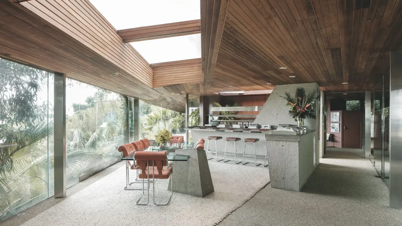 Interno della Sheats Goldstein Residence con soffitto in legno, ampie vetrate, zona pranzo con tavolo in vetro e sedie in pelle arancione, e un bancone bar in cemento con sgabelli.