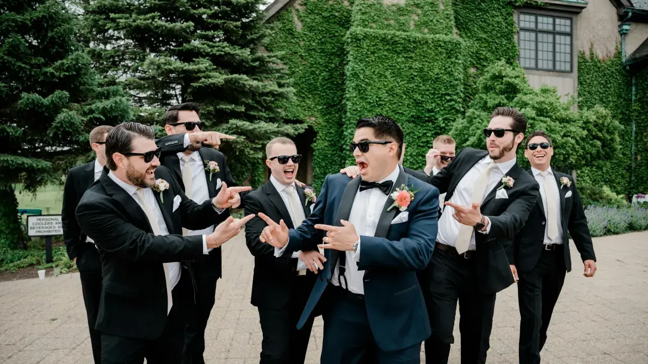 Groomsmen in black suits and cream ties, with the groom in a navy suit, all sporting sunglasses and a playful mood. Do groomsmen match the groom? Not exactly, but they're all dressed to impress!