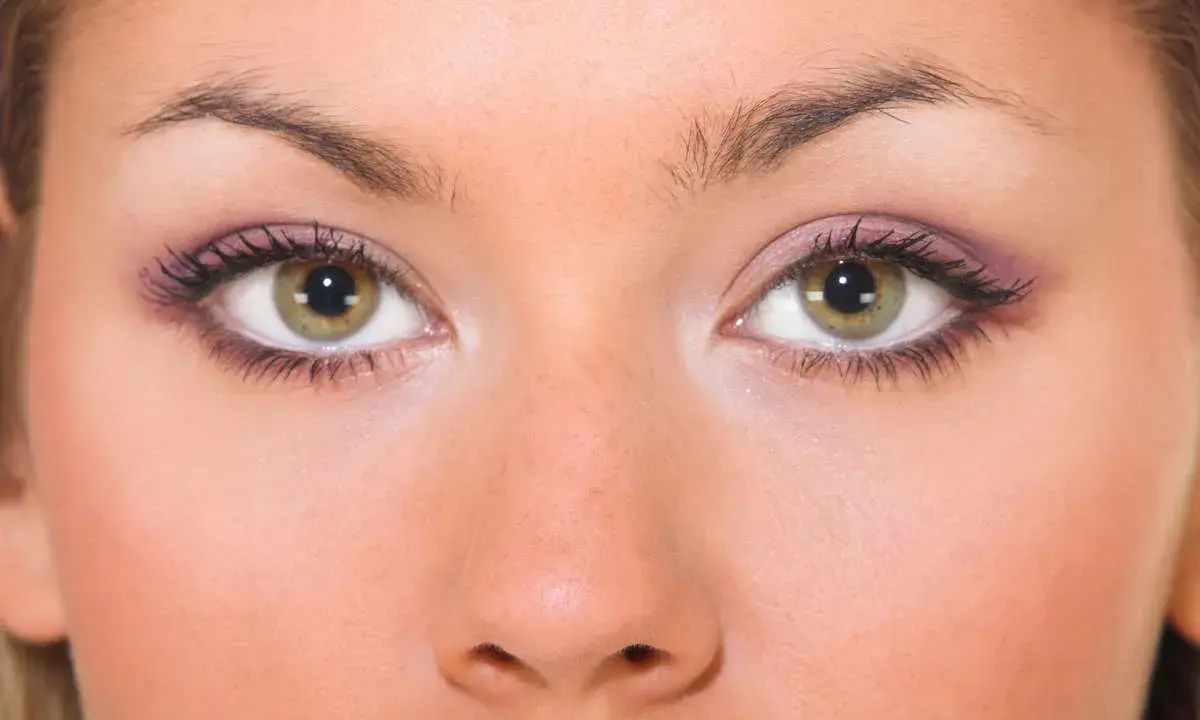 Primo piano di un viso femminile con occhi piccoli e verdi, ciglia lunghe e trucco viola sulle palpebre.