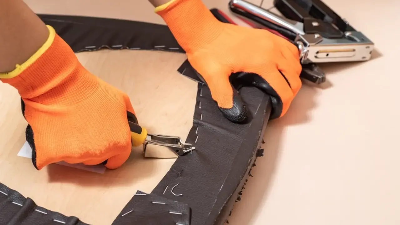 Manos con guantes naranjas usando una grapadora para tapizar una silla.