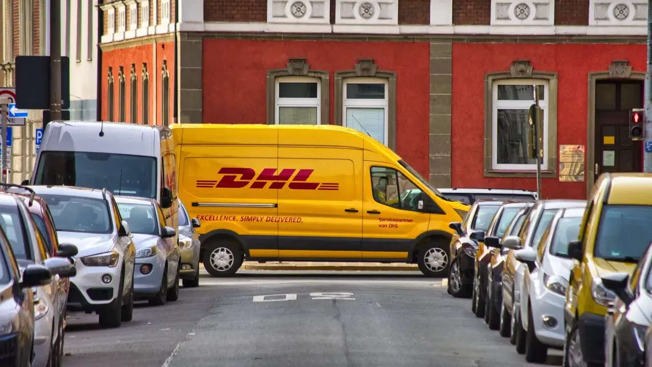 Żółta furgonetka DHL stoi zaparkowana na ulicy, obok innych samochodów. To miejsce, do którego pracują kurierzy DHL, by dostarczać paczki.