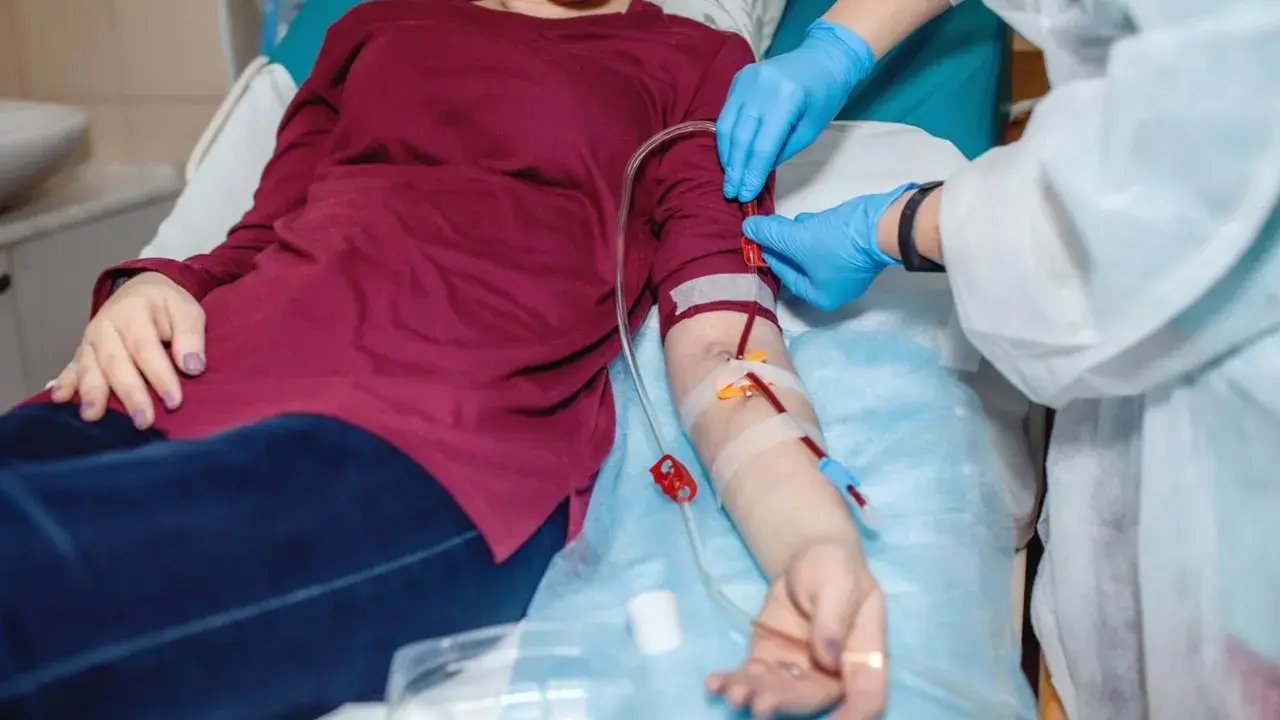 Pacjentka leży, a pielęgniarka w rękawiczkach podłącza przetokę do dializ. Krew płynie przez rurki.