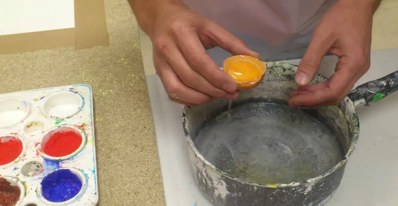 Manos vierten una yema de huevo en un recipiente para preparar una pintura al temple. Paleta con pigmentos de colores al lado.