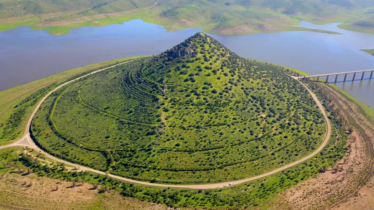 El **cerro Masatrigo**, cubierto de vegetación, se alza majestuoso junto a un embalse. Una carretera serpentea a su alrededor, conectando con un puente.