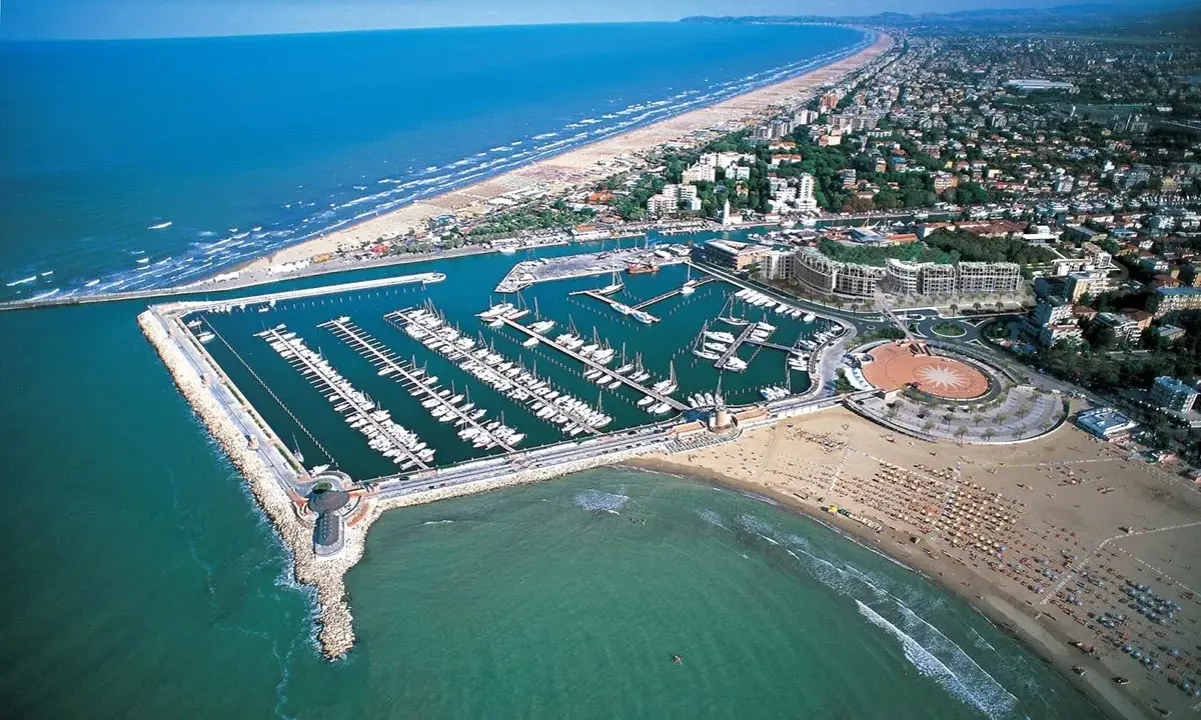 La darsena di Rimini, con le sue barche ormeggiate, si estende lungo la costa adriatica, tra la spiaggia affollata e il mare azzurro.