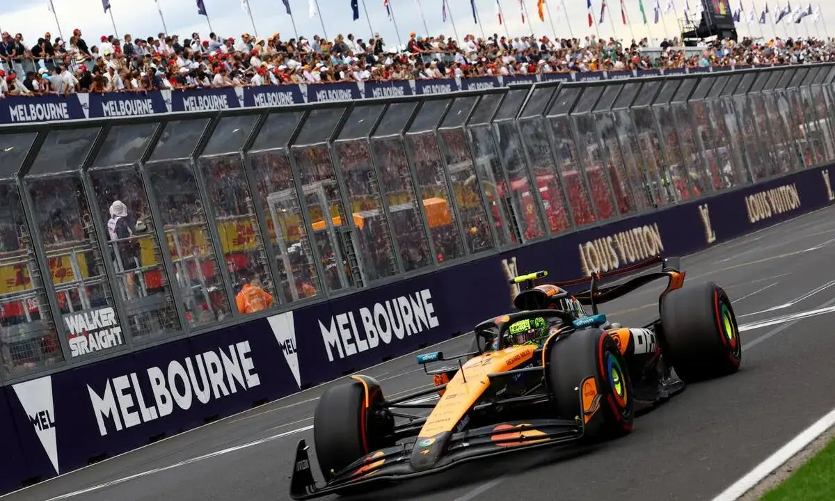 Un coche de Fórmula 1 naranja y negro acelera en Melbourne. Los aficionados vitorean. ¡Qué ganas de que empiece la Fórmula 1 2026!