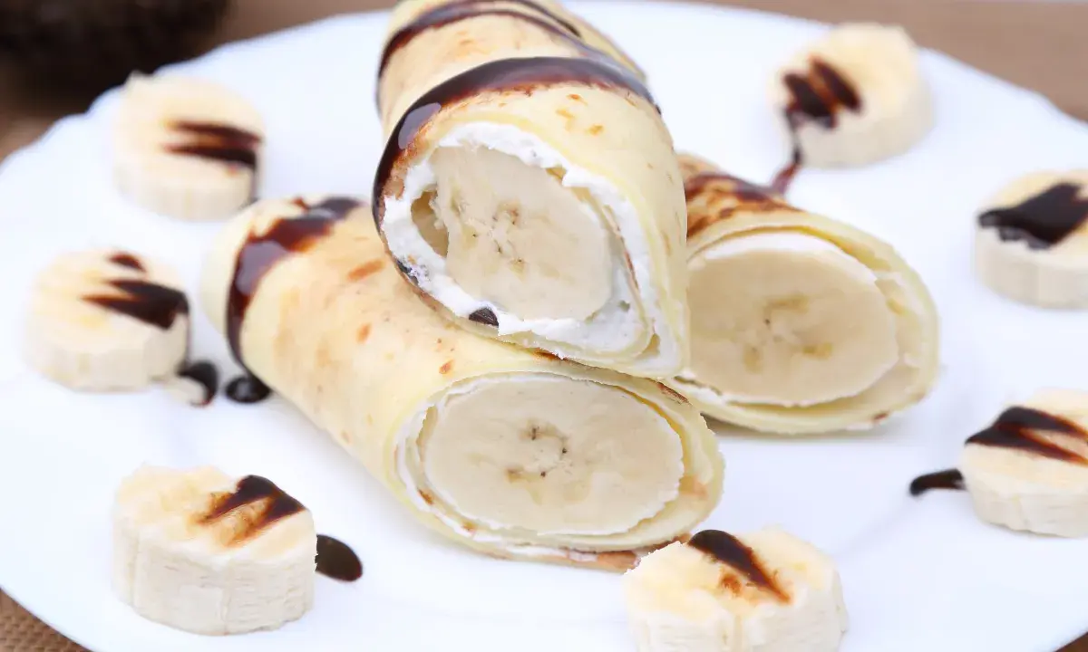 Rolowane naleśniki z bananem i kremem, polane czekoladą. Na talerzu ułożone są też kawałki banana z polewą.