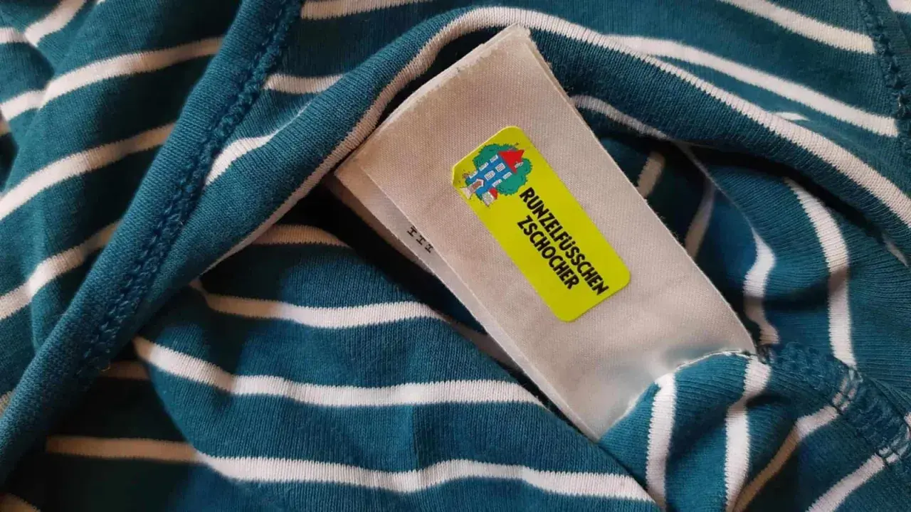 Namensetiketten Kita Erfahrungen: Ein gestreiftes Shirt mit einem gelben Aufkleber "Runzelfüßchen Zschocher" und einem Hausmotiv.