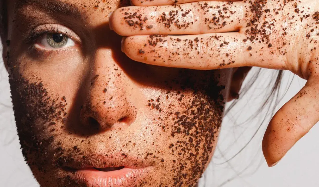 Visage couvert de grains d'un exfoliant chimique naturel, une main cache un œil.