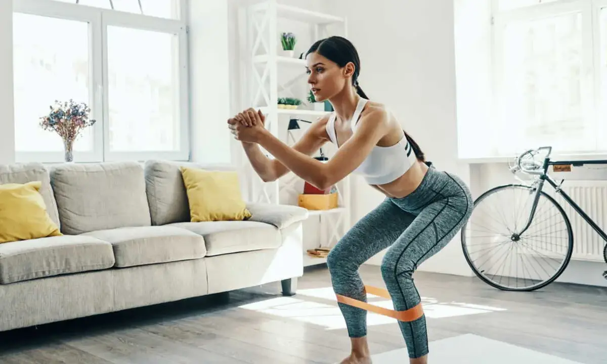 Femme faisant des squats avec une bande de résistance pour affiner ses jambes, dans un salon lumineux.