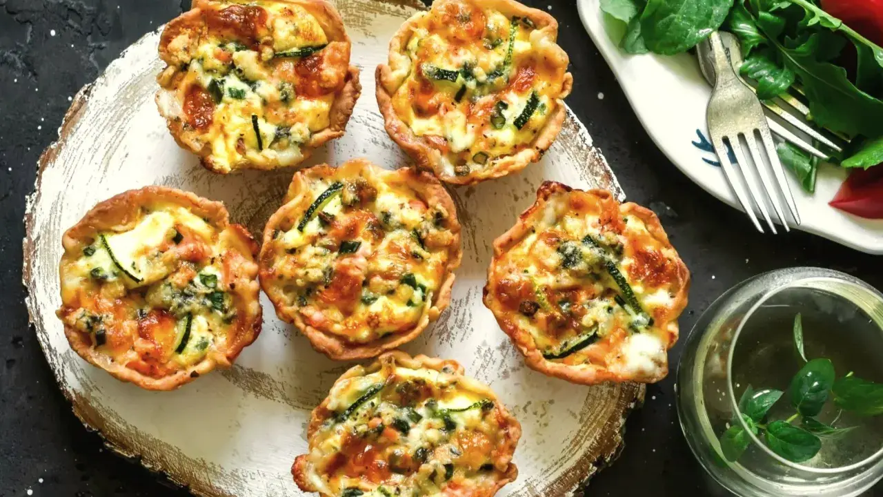 Sześć apetycznych, kruche babeczki na słono z cukinią i serem, podane na drewnianej desce. Obok sałatka z pomidorami i widelcem.