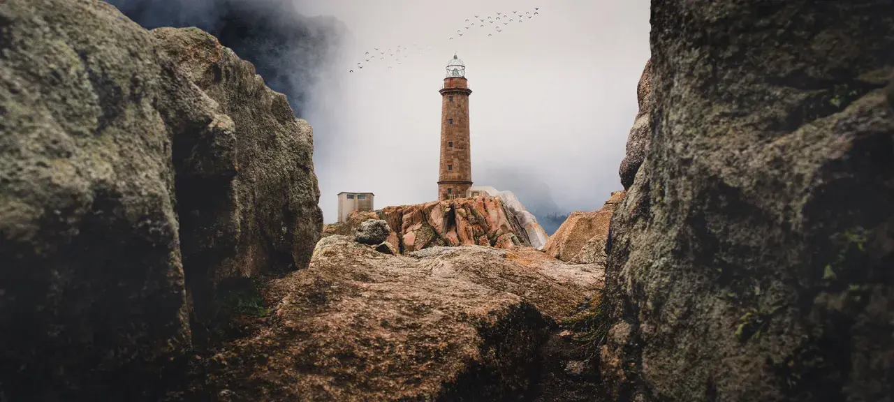 Leuchtturm auf felsiger Küste der Costa da Morte, Vögel ziehen über neblige Berge.