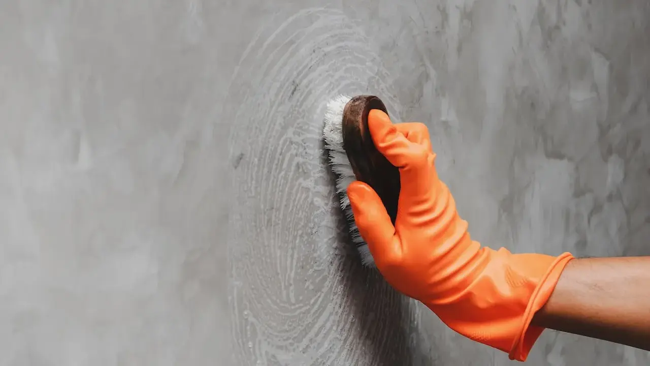 Mano con guante naranja limpiando pared gris con cepillo, aprendiendo como limpiar las paredes de color sin dañar la pintura.