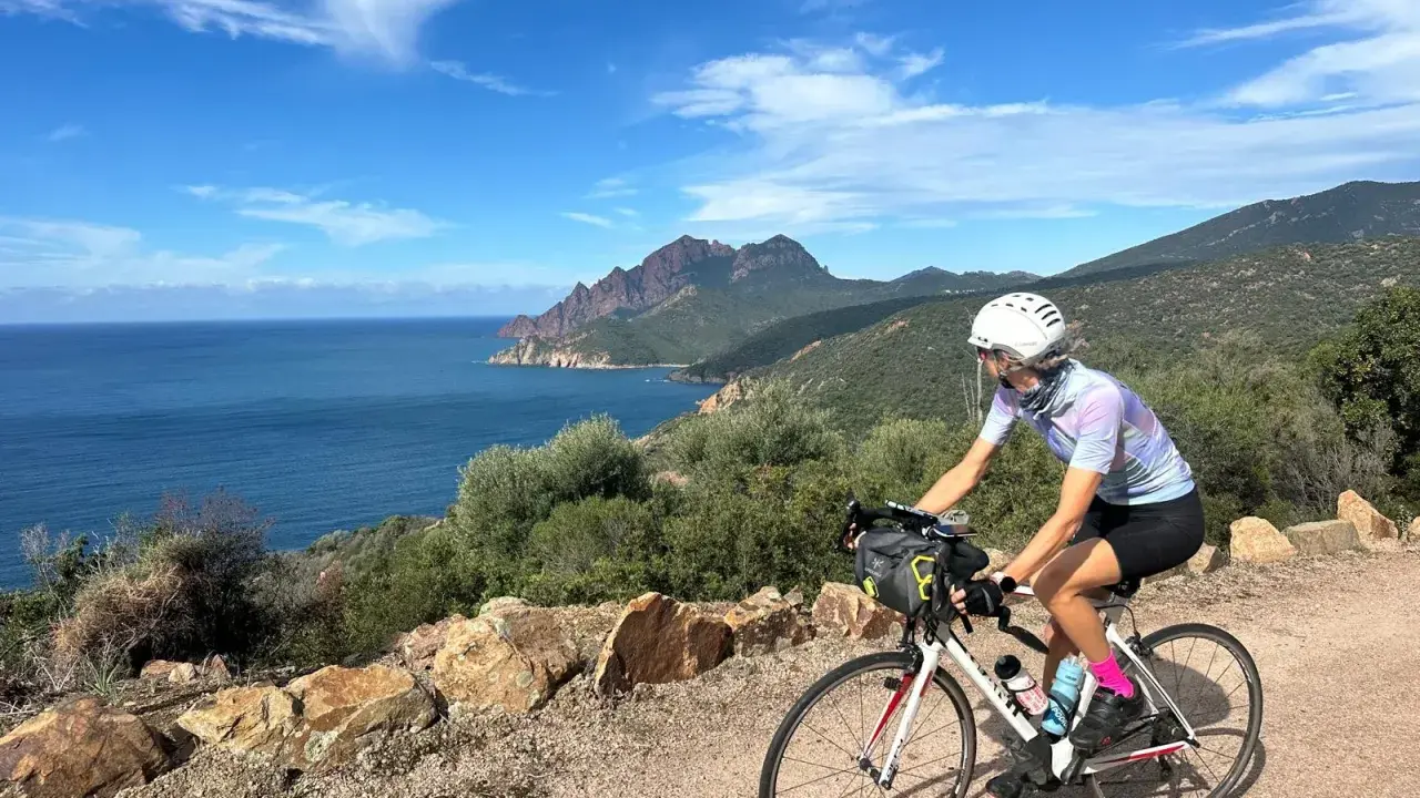 Ciclista pedala lungo la costa rocciosa della Corsica, con il mare blu e montagne sullo sfondo. Un'avventura in bici indimenticabile.
