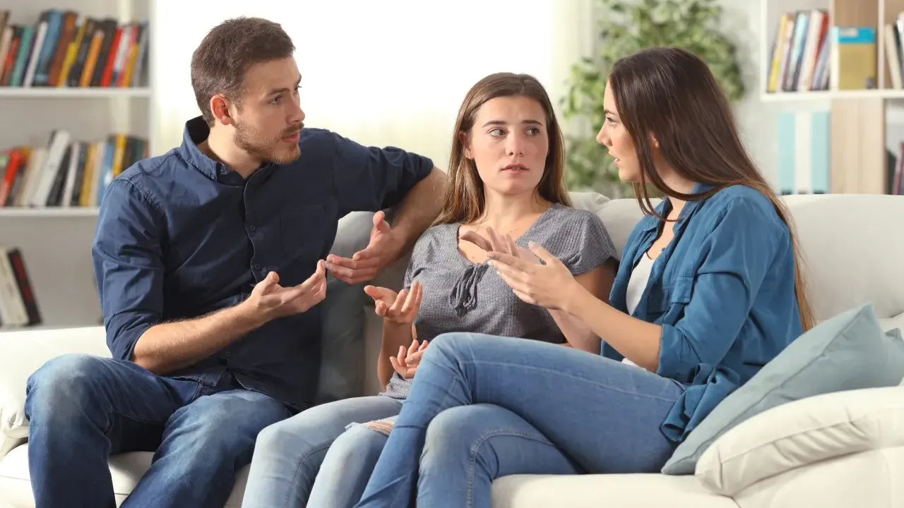 Tres personas sentadas en un sofá blanco discuten animadamente, gesticulando con las manos, en un intento por resolver conflictos.