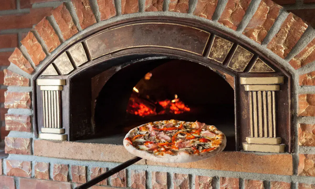 Une pizza garnie est sur le point d'entrer dans un four à pizza extérieur en briques rouges, avec des flammes visibles à l'intérieur.