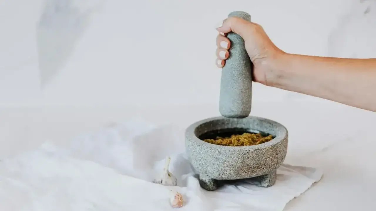 Dłoń uciera w moździerzu aromatyczne pesto z bazylii. Obok leżą ząbki czosnku.