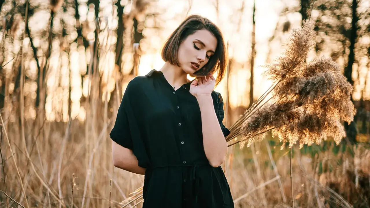 Młoda kobieta w czarnej sukience pozuje na tle zachodzącego słońca i trzcin. Idealna modelka do nauki kurs fotografii portretowej.