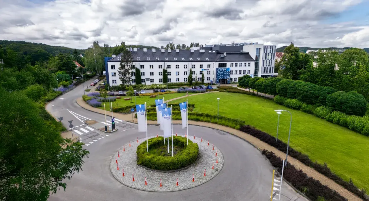Nowoczesny budynek WSB Gdańsk otoczony zielenią. Na rondzie powiewają białe flagi z logo uczelni.