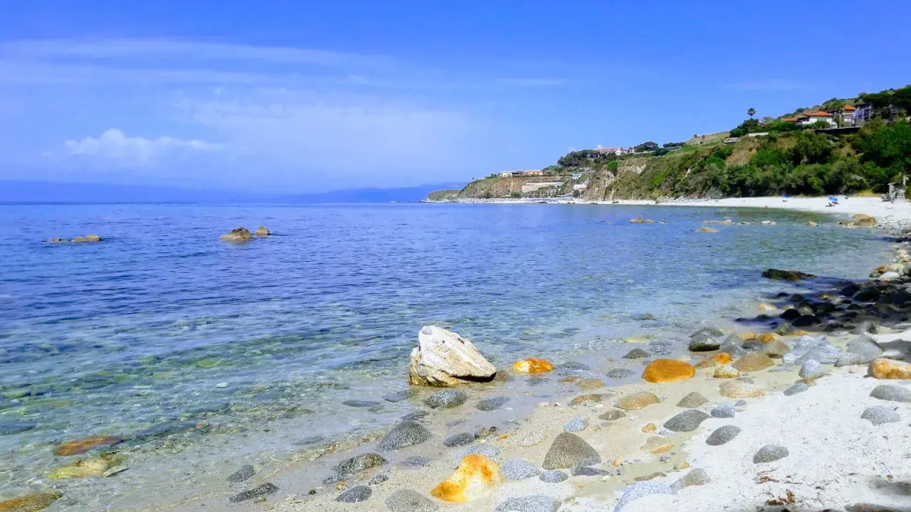 Spiaggia di Tropea con la torre Sant'Irene in lontananza, mare cristallino e rocce sulla riva.