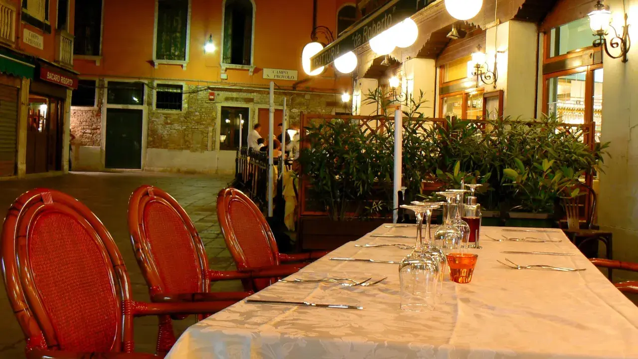 Table dressée devant la trattoria "da Roberto" à Venise, avec des chaises rouges et des verres à vin.