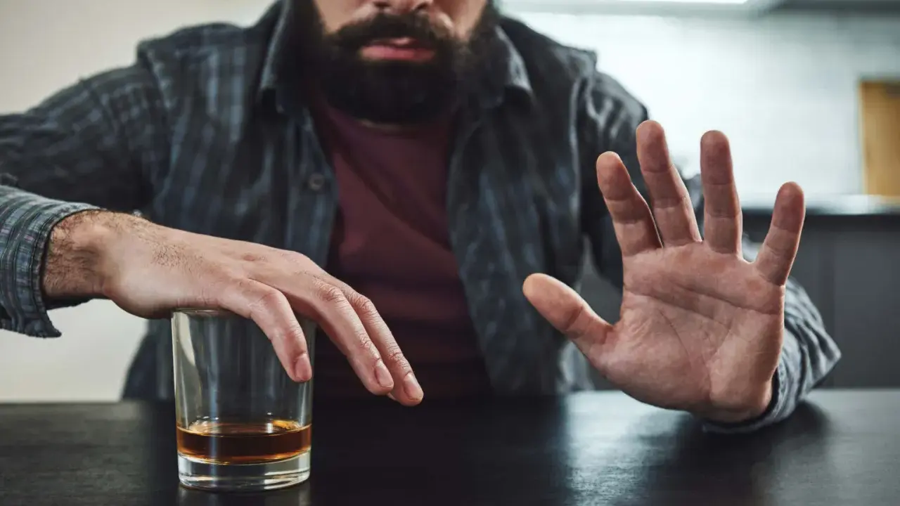 Mężczyzna z zarostem odpycha szklankę z alkoholem, walcząc z chorobą alkoholową. Gest dłoni symbolizuje odmowę i chęć zmiany.