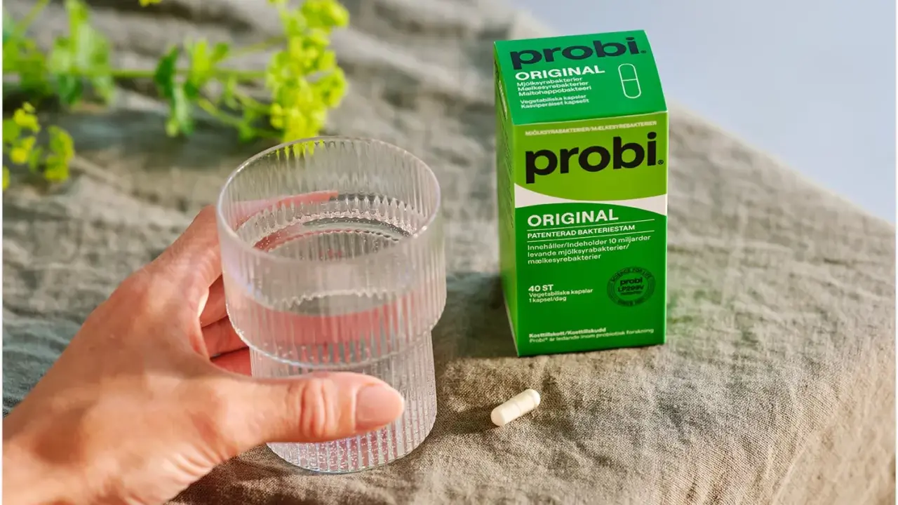 En hand håller ett glas vatten bredvid en förpackning med probi. En kapsel ligger på bordet, redo att tas för att undvika probi mage biverkningar.